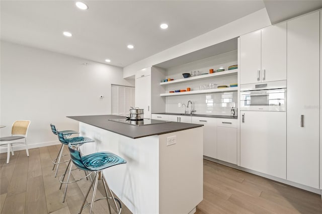 kitchen with black electric cooktop, dark countertops, open shelves, light wood finished floors, and a kitchen breakfast bar