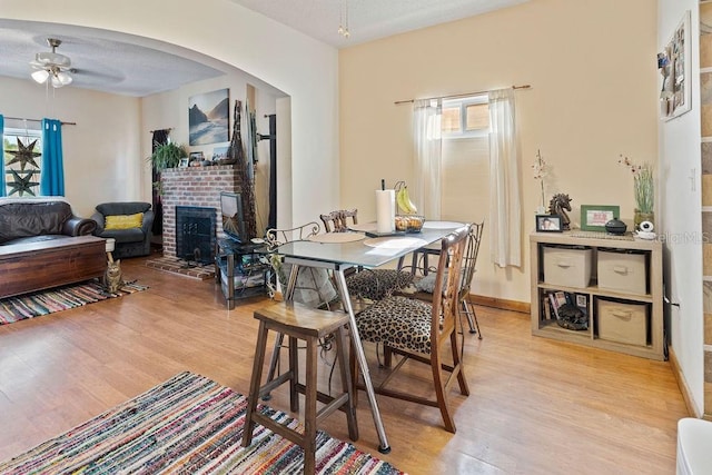 dining room with light wood-style flooring, a ceiling fan, arched walkways, and a brick fireplace
