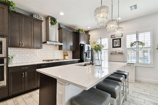 kitchen with wall chimney exhaust hood, a sink, appliances with stainless steel finishes, a chandelier, and tasteful backsplash