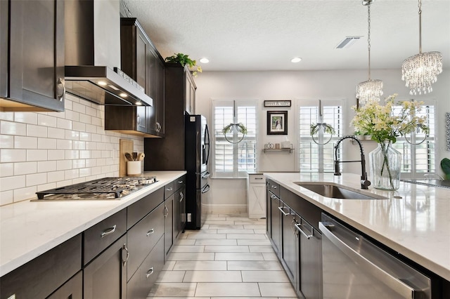 kitchen featuring wall chimney range hood, a sink, stainless steel appliances, a chandelier, and dark brown cabinetry