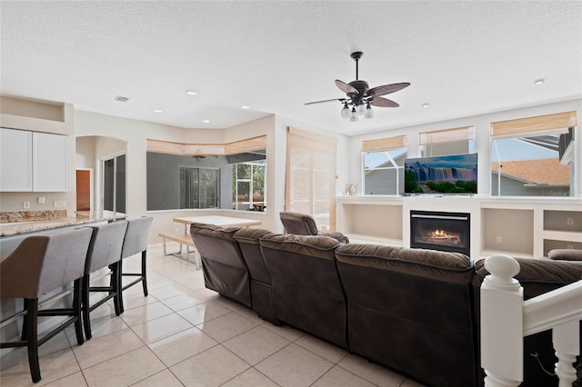 living area with light tile patterned floors, a glass covered fireplace, a ceiling fan, a textured ceiling, and recessed lighting