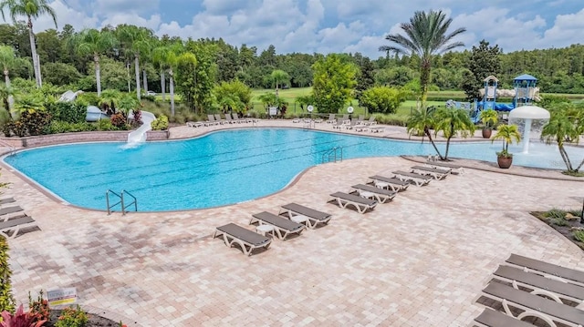 community pool with a water slide, a patio, view of wooded area, and fence