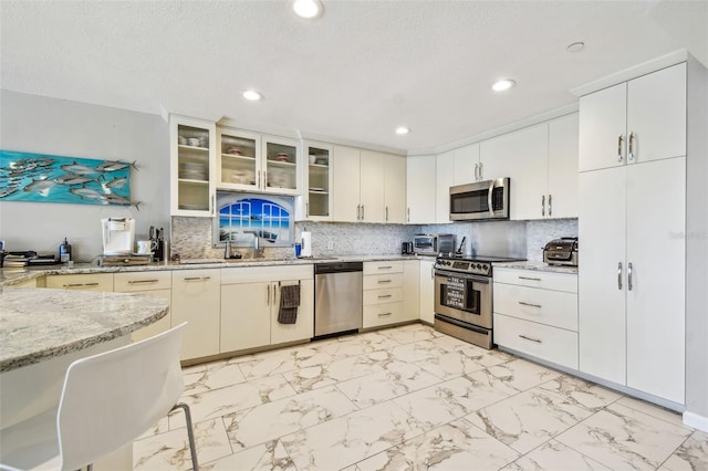 kitchen featuring appliances with stainless steel finishes, light stone countertops, light marble finish flooring, glass insert cabinets, and recessed lighting