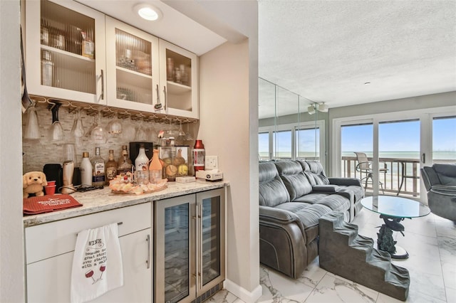 indoor dry bar featuring light marble finish floors, beverage cooler, decorative backsplash, a water view, and a textured ceiling