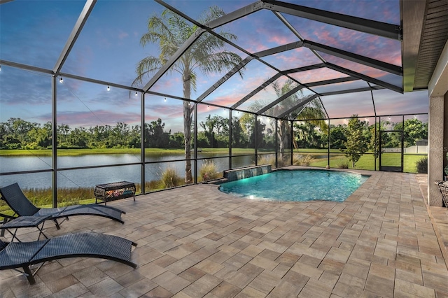 pool at dusk with a patio area, a water view, an outdoor pool, glass enclosure, and view of wooded area