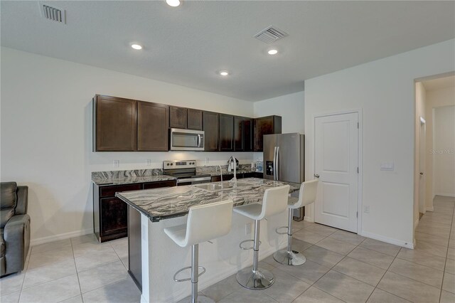 33062 Frosted Clover Way, Wesley Chapel, FL 33545 - kitchen with stainless steel appliances, a kitchen bar, light stone counters, dark brown cabinets, and a kitchen island with sink