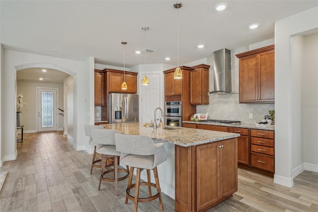 19183 Coastal Shore Ter, Land O Lakes, FL 34638 - kitchen featuring stainless steel appliances, wall chimney exhaust hood, arched walkways, light wood finished floors, and brown cabinetry