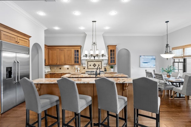 7713 Still Lakes Dr, Odessa, FL 33556 - kitchen with stainless steel built in fridge, arched walkways, brown cabinetry, an island with sink, and decorative backsplash