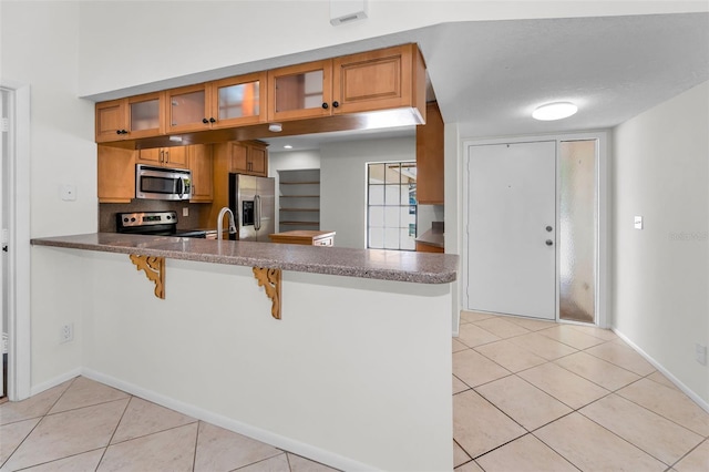 3041 Oak View Dr, Palm Harbor, FL 34684 - kitchen with stainless steel appliances, a peninsula, light tile patterned flooring, brown cabinetry, and glass insert cabinets