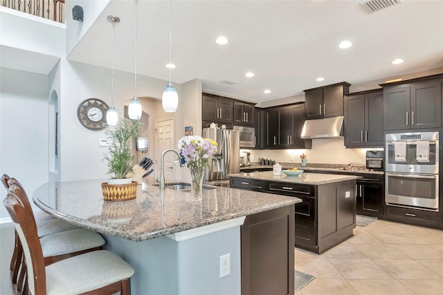 26972 Evergreen Chase Dr, Wesley Chapel, FL 33544 - kitchen featuring an island with sink, under cabinet range hood, stainless steel appliances, light stone counters, and light tile patterned floors