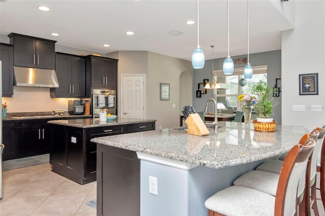 26972 Evergreen Chase Dr, Wesley Chapel, FL 33544 - kitchen featuring under cabinet range hood, a kitchen island with sink, light stone counters, light tile patterned flooring, and a kitchen bar
