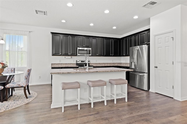 11434 Leland Groves Dr, Riverview, FL 33579 - kitchen featuring appliances with stainless steel finishes, dark cabinets, a center island with sink, a kitchen breakfast bar, and light stone countertops