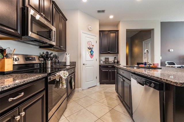 11711 Winterset Cove Dr, Riverview, FL 33579 - kitchen featuring stainless steel appliances, light tile patterned floors, dark stone countertops, a kitchen island with sink, and dark brown cabinetry