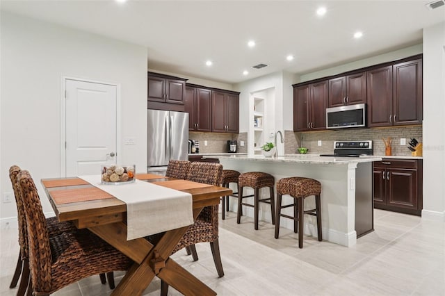5808 Grand Sonata Ave, Lutz, FL 33558 - kitchen with stainless steel appliances, a kitchen island with sink, dark brown cabinetry, tasteful backsplash, and a breakfast bar