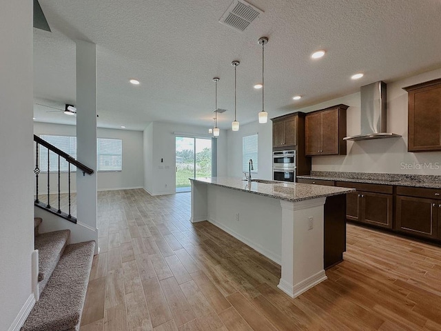 38120 Shale Stone Ct, Zephyrhills, FL 33540 - kitchen with wall chimney exhaust hood, light stone counters, a textured ceiling, an island with sink, and light wood-style floors