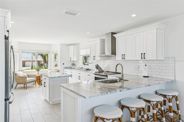 311 22nd St, Belleair Beach, FL 33786 - kitchen featuring wall chimney range hood, a peninsula, backsplash, light stone countertops, and white cabinetry