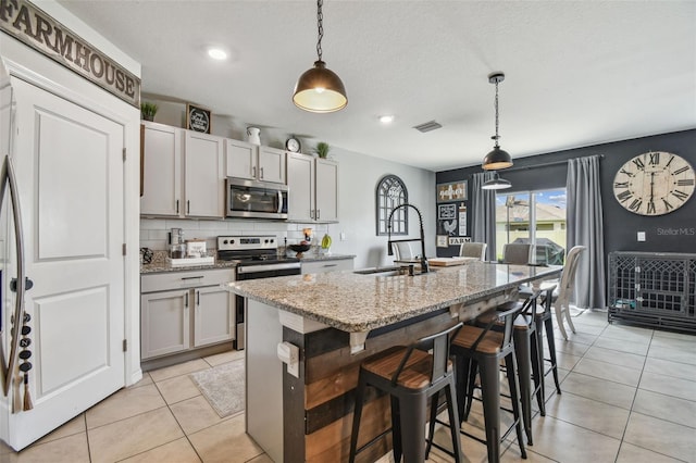 33678 Jasper Stone Dr, Wesley Chapel, FL 33543 - kitchen featuring appliances with stainless steel finishes, light tile patterned floors, a kitchen island with sink, light stone counters, and hanging light fixtures