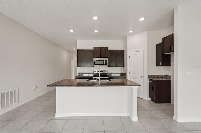 13008 Majestic Garden Ln, Riverview, FL 33579 - kitchen with stainless steel appliances, light tile patterned flooring, an island with sink, dark stone counters, and dark brown cabinets