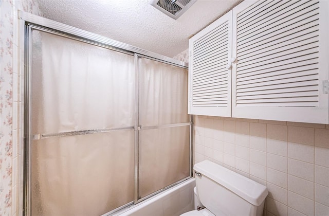 bathroom featuring tile walls, shower / bath combination with glass door, toilet, and a textured ceiling