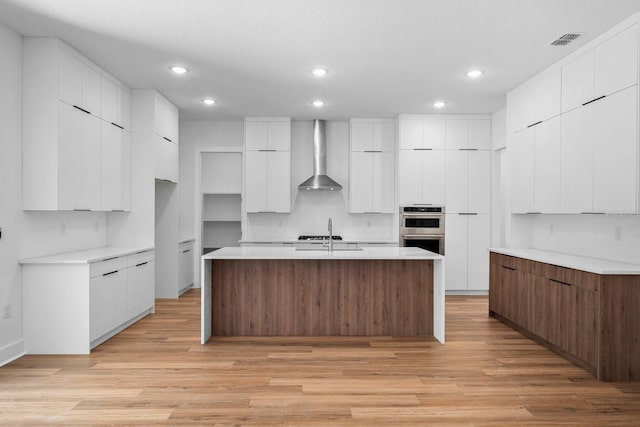 kitchen with white cabinetry, light wood-type flooring, wall chimney range hood, and a kitchen island with sink