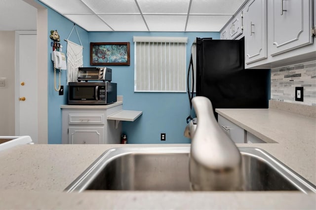 kitchen with a paneled ceiling, backsplash, black refrigerator, and white cabinetry