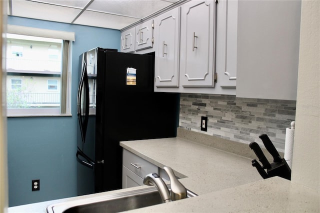 kitchen with sink, backsplash, white cabinets, black fridge, and a drop ceiling