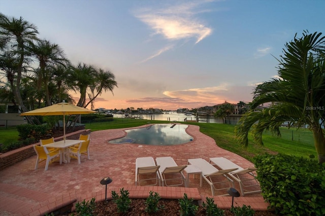 pool at dusk featuring a patio area, a water view, and a lawn