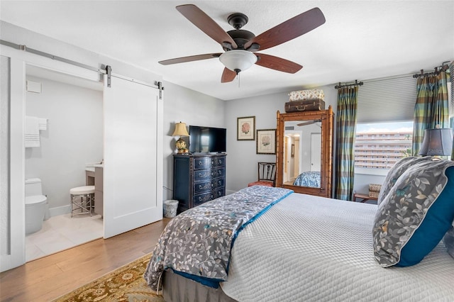 bedroom with a barn door, wood-type flooring, ensuite bath, and ceiling fan