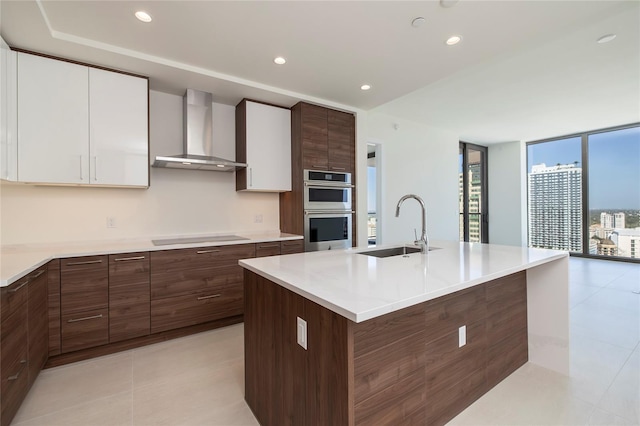 kitchen with sink, stainless steel double oven, a center island with sink, wall chimney exhaust hood, and white cabinetry