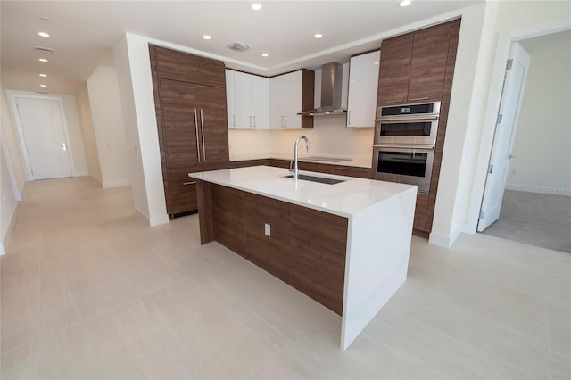 kitchen featuring stainless steel double oven, an island with sink, light tile floors, white cabinets, and wall chimney exhaust hood