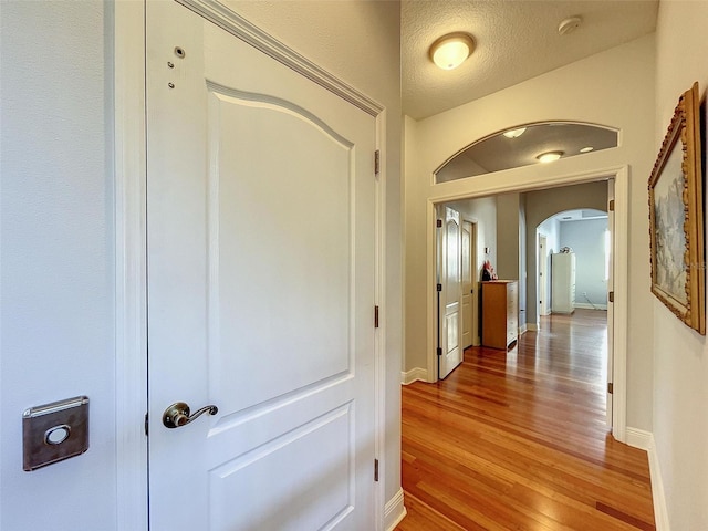 hallway featuring arched walkways, light wood finished floors, a textured ceiling, and baseboards