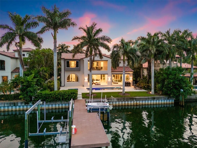 back of house at dusk with boat lift, stucco siding, a water view, a patio area, and a balcony