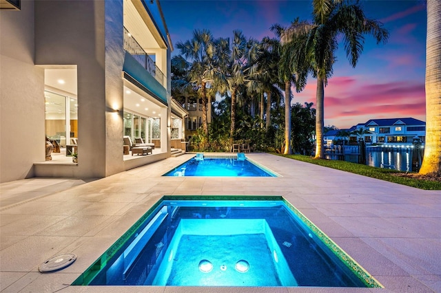 pool at dusk featuring a water view, a patio area, and an in ground hot tub