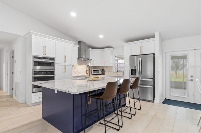 kitchen featuring appliances with stainless steel finishes, light wood-type flooring, wall chimney exhaust hood, a kitchen island with sink, and white cabinets