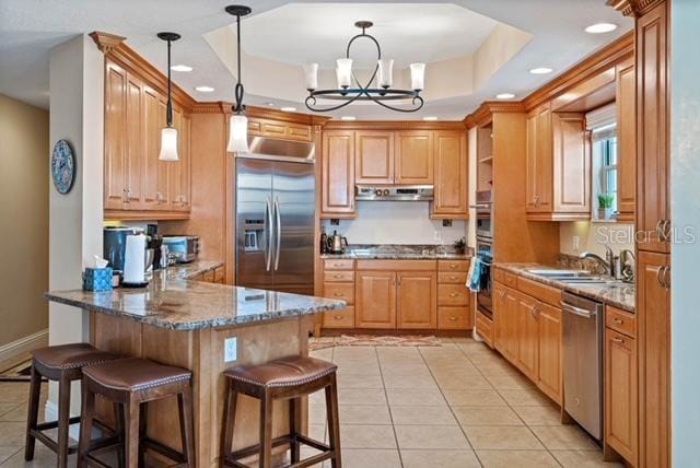 kitchen with light tile patterned floors, ventilation hood, a tray ceiling, stainless steel appliances, and decorative light fixtures