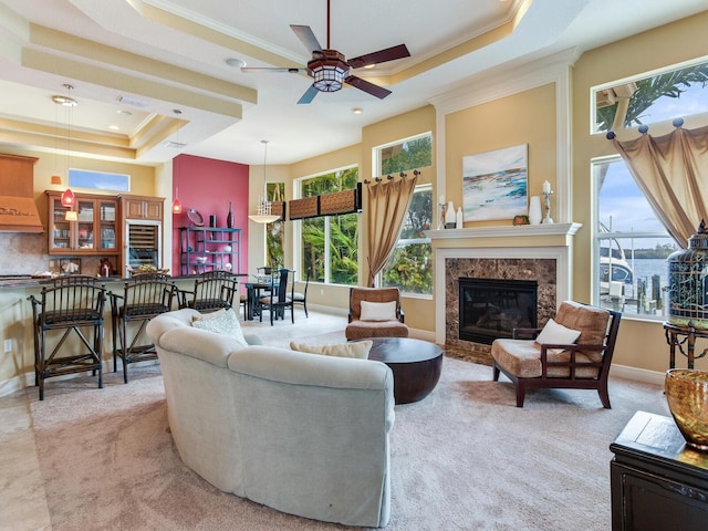 carpeted living room with a high end fireplace, a raised ceiling, ceiling fan, and ornamental molding