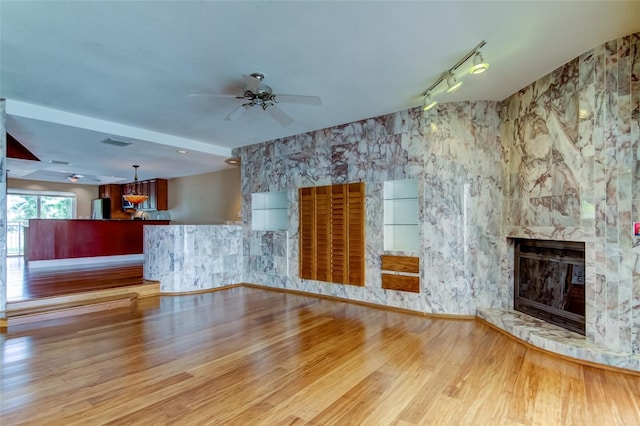 unfurnished living room featuring rail lighting, a stone fireplace, ceiling fan, and light wood-type flooring