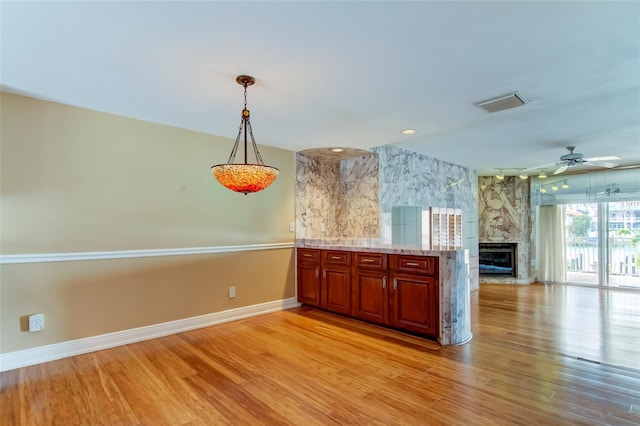 kitchen with ceiling fan, light stone counters, hanging light fixtures, a stone fireplace, and light wood-type flooring