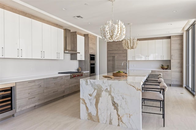 kitchen featuring a kitchen island with sink, light stone counters, wall chimney range hood, an inviting chandelier, and white cabinetry