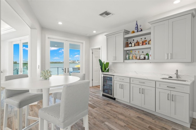 bar featuring light stone counters, beverage cooler, a water view, light hardwood / wood-style flooring, and gray cabinets