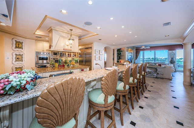 kitchen featuring ceiling fan, a breakfast bar, stainless steel appliances, decorative columns, and a tray ceiling