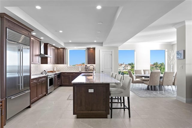 kitchen with light stone counters, stove, light tile flooring, stainless steel built in refrigerator, and wall chimney exhaust hood