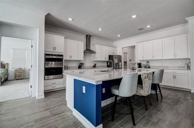 kitchen with a breakfast bar, white cabinets, double oven, wall chimney range hood, and a center island with sink