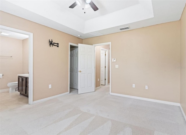 unfurnished bedroom with light carpet, ceiling fan, ensuite bathroom, and a raised ceiling