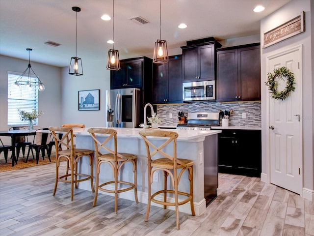 kitchen featuring appliances with stainless steel finishes, decorative light fixtures, light hardwood / wood-style flooring, a breakfast bar area, and an island with sink