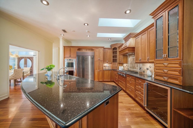 kitchen featuring an island with sink, appliances with stainless steel finishes, light hardwood / wood-style floors, beverage cooler, and tasteful backsplash