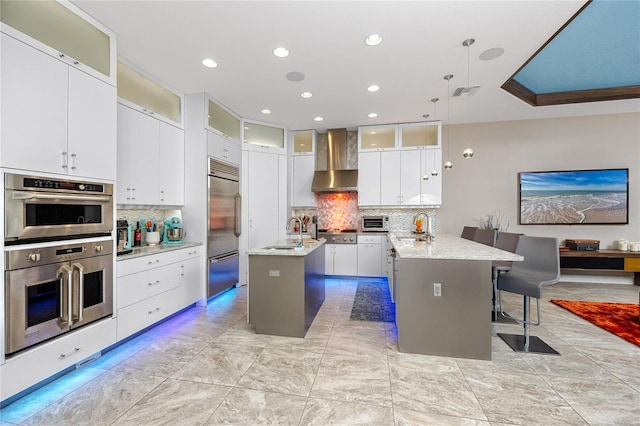 kitchen featuring appliances with stainless steel finishes, decorative light fixtures, wall chimney range hood, white cabinetry, and an island with sink