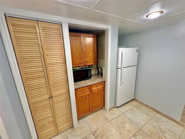 kitchen with baseboards, black microwave, light stone counters, freestanding refrigerator, and brown cabinetry