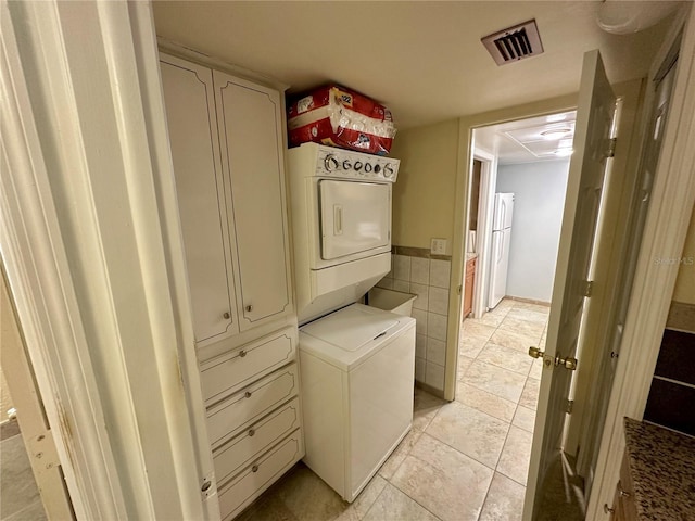 clothes washing area featuring visible vents, light tile patterned floors, stacked washer and clothes dryer, cabinet space, and tile walls