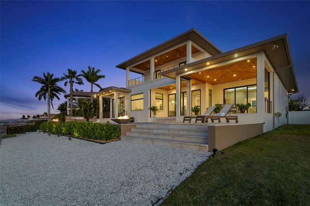 back house at dusk featuring a balcony, a patio, and a lawn